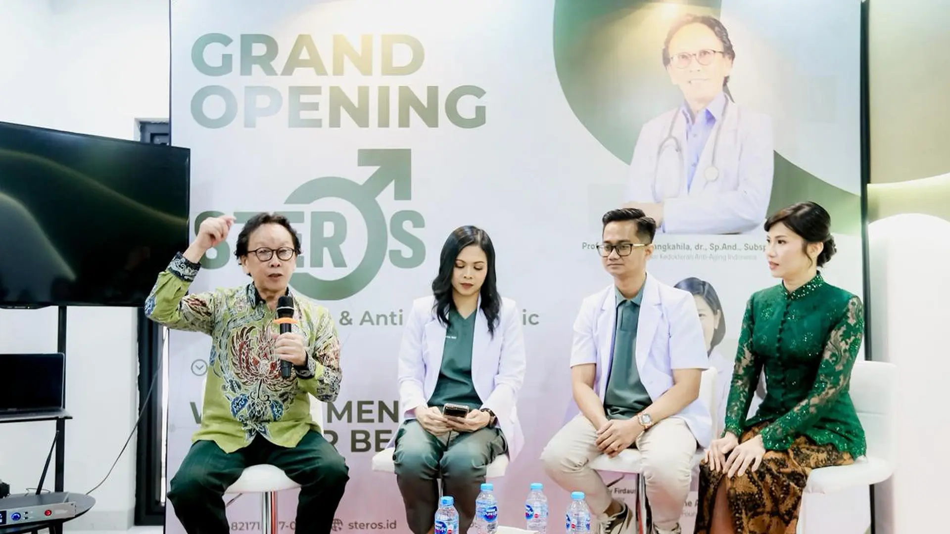 Four people seated at a grand opening event, one man is speaking into a microphone while others listen; backdrop shows 'Grand Opening Steros' with medical imagery.