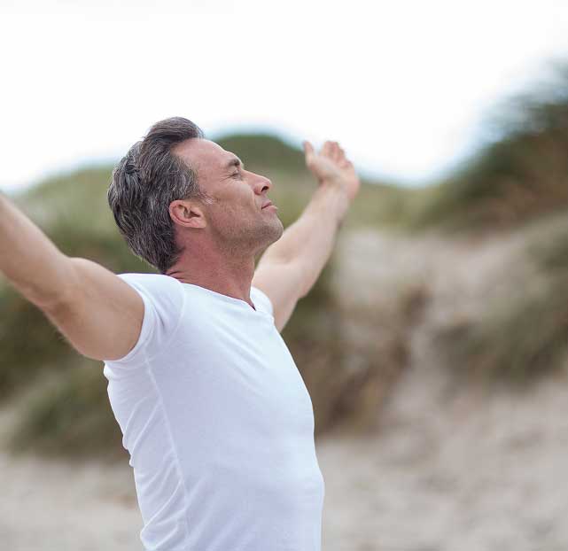 Middle-aged man in a white shirt standing outdoors with arms wide open and eyes closed, enjoying fresh air.